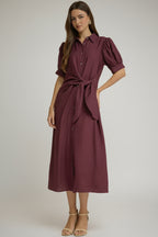 A woman wearinga midi-length, short-sleeved button-up dress in a wine shade. The dress features a side tie waist, puffed short sleeves, and a relaxed skirt, styled with beige strappy high-heeled sandals against a neutral gray studio background.
