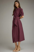 A woman wearinga midi-length, short-sleeved button-up dress in a wine shade. The dress features a side tie waist, puffed short sleeves, and a relaxed skirt, styled with beige strappy high-heeled sandals against a neutral gray studio background.