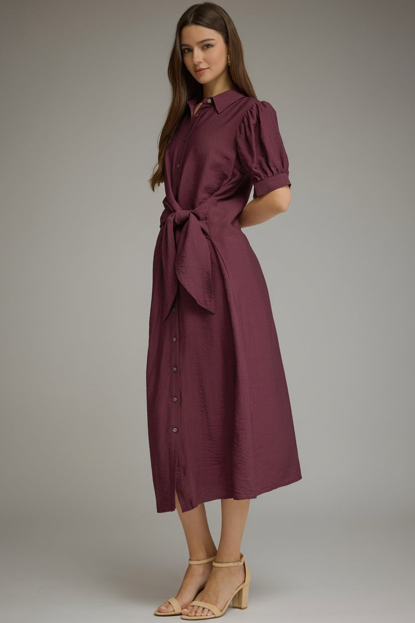 A woman wearinga midi-length, short-sleeved button-up dress in a wine shade. The dress features a side tie waist, puffed short sleeves, and a relaxed skirt, styled with beige strappy high-heeled sandals against a neutral gray studio background.