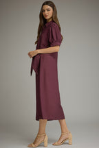 A woman wearinga midi-length, short-sleeved button-up dress in a wine shade. The dress features a side tie waist, puffed short sleeves, and a relaxed skirt, styled with beige strappy high-heeled sandals against a neutral gray studio background.