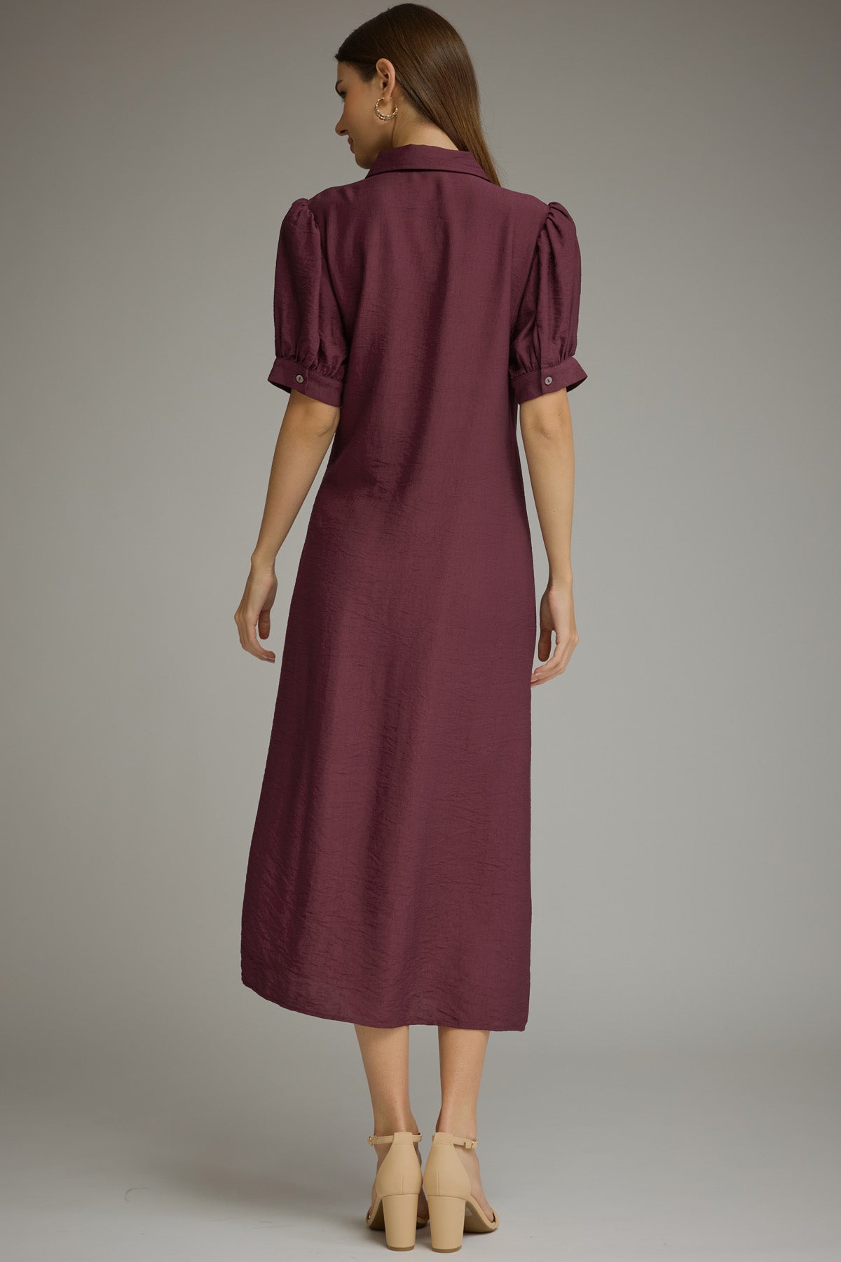 Back view of a woman wearinga midi-length, short-sleeved button-up dress in a wine shade. The dress features a side tie waist, puffed short sleeves, and a relaxed skirt, styled with beige strappy high-heeled sandals against a neutral gray studio background.