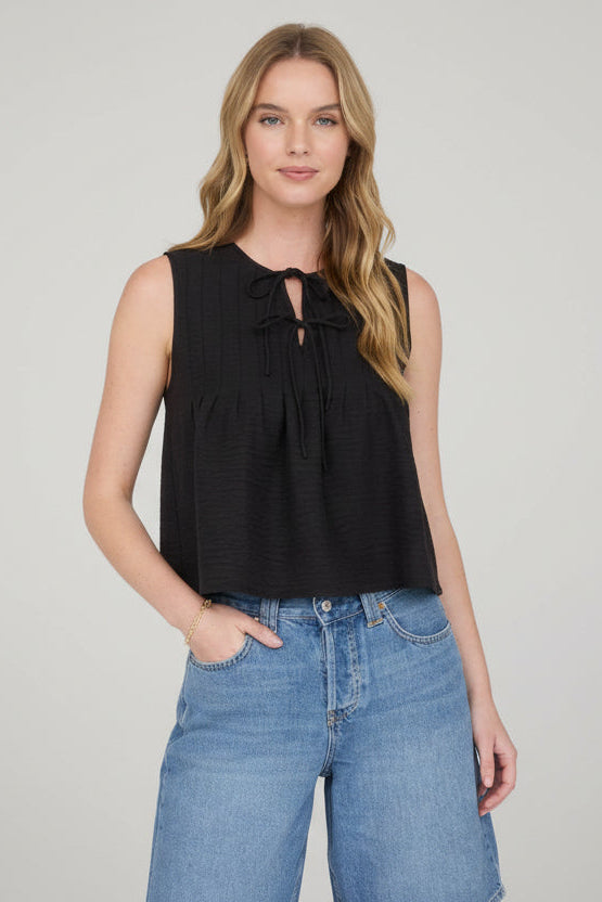 Woman wearing sleeveless black pintuck detail front tie top with jeans on a detail background