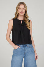 Woman wearing sleeveless black pintuck detail front tie top with jeans on a detail background