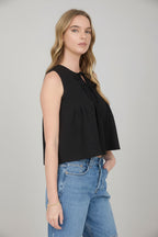 Woman wearing sleeveless black pintuck detail front tie top with jean shorts on a detailed background