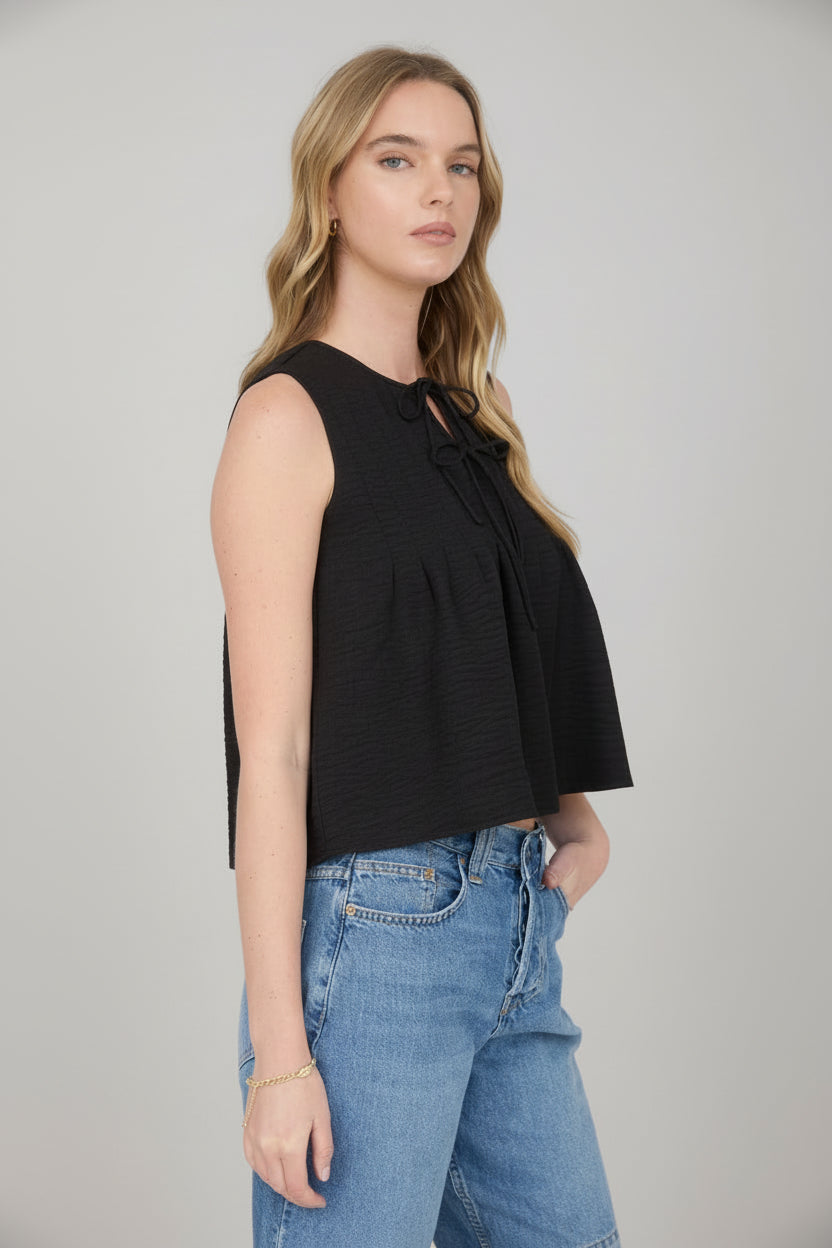 Woman wearing sleeveless black pintuck detail front tie top with jean shorts on a detailed background