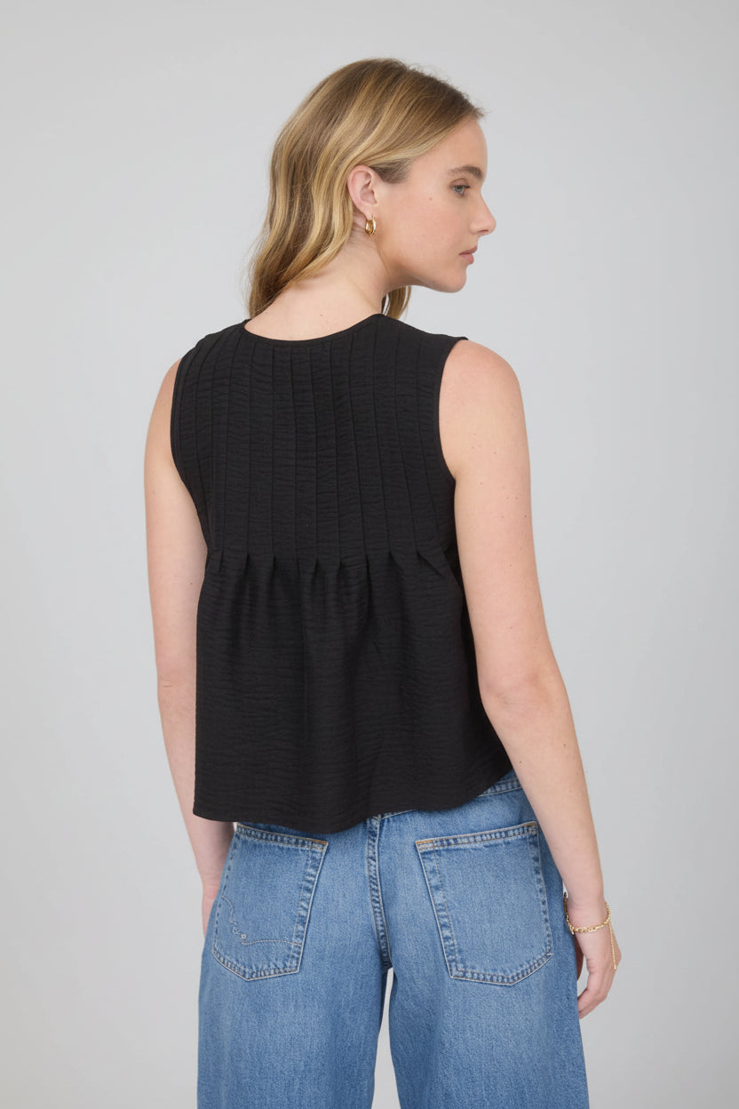 Back view of a woman wearing a sleeveless black pintuck detail tie top with jeans on a detail background