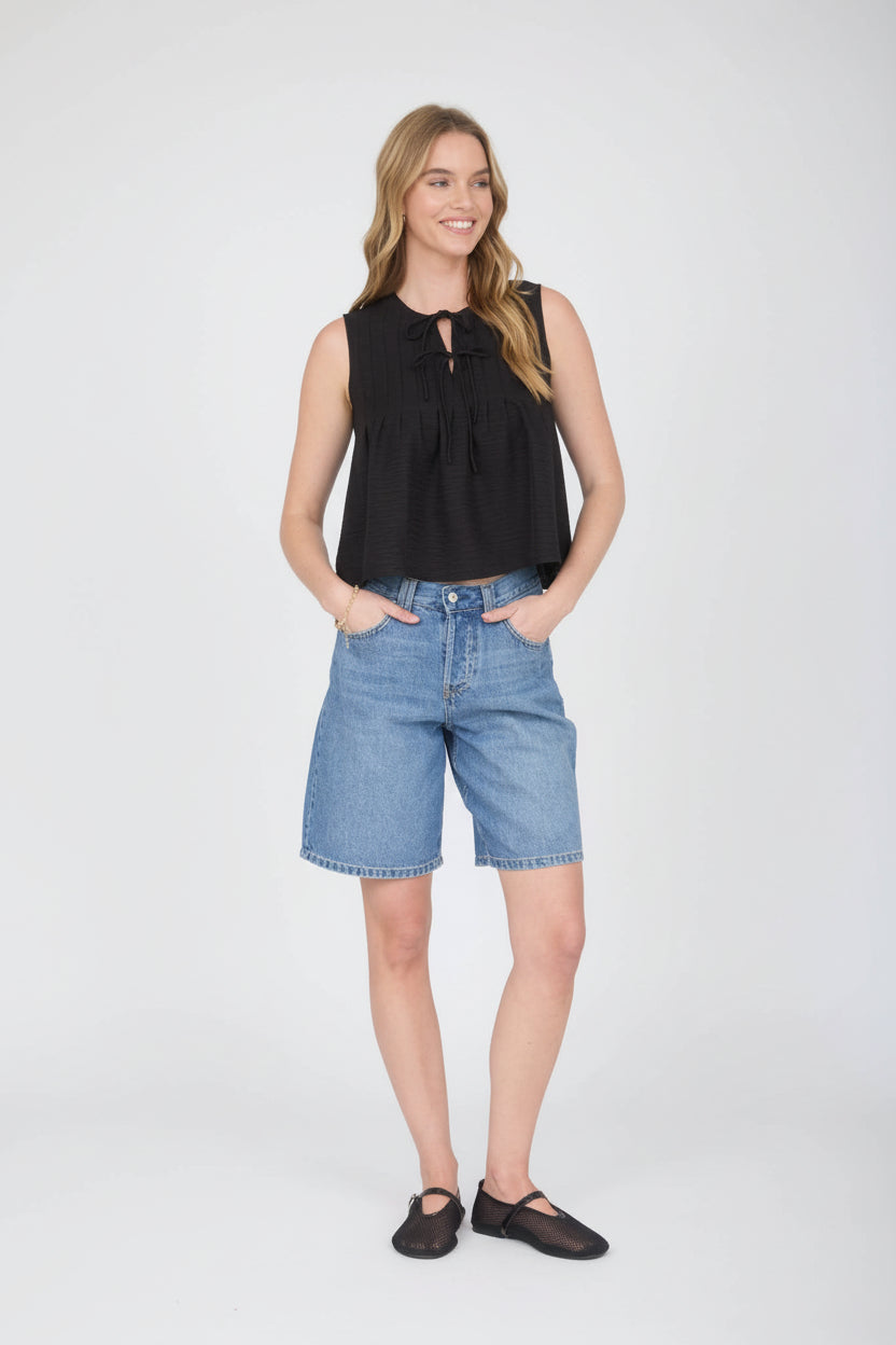 Woman wearing sleeveless black pintuck detail front tie top with jean shorts on a detailed background