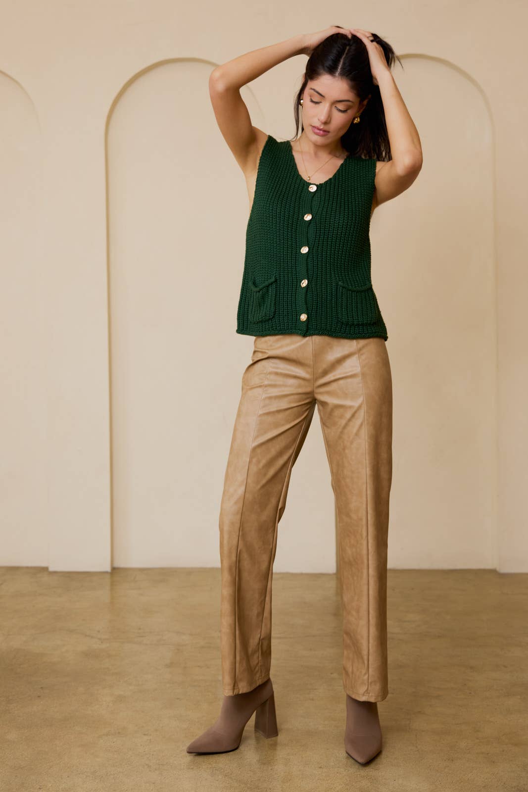 Woman wearing a green knitted vest with gold buttons and gold pants against white background.