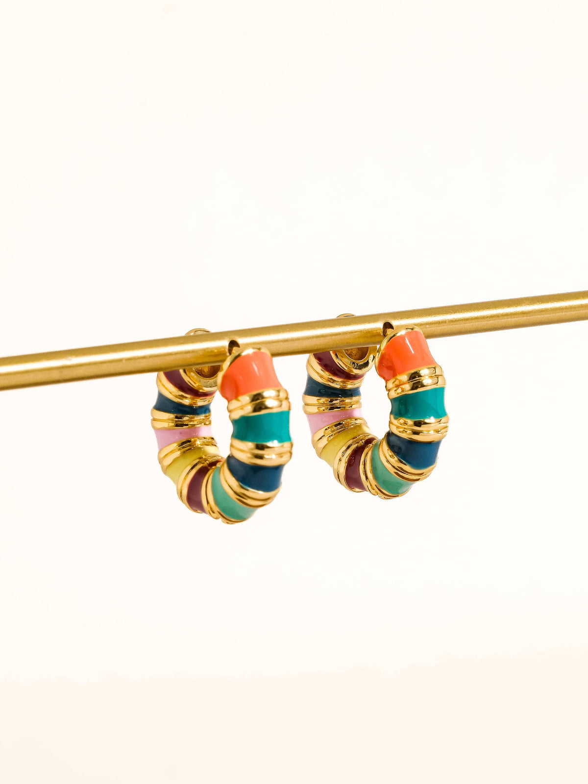 A pair of gold hoop earrings with multi-colored striped enamel bands in coral, teal, purple, and pink, hanging from a gold display rod against a plain white background.